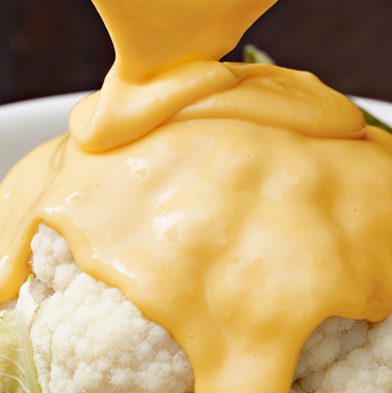 Creamy yellow cheese sauce being poured on to cauliflower floret in a white ceramic bowl